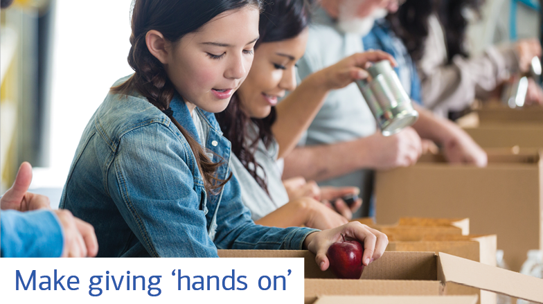 A young girl puts an apple in a box while other people put various items into boxes. Text reads: Make giving ‘hands on’